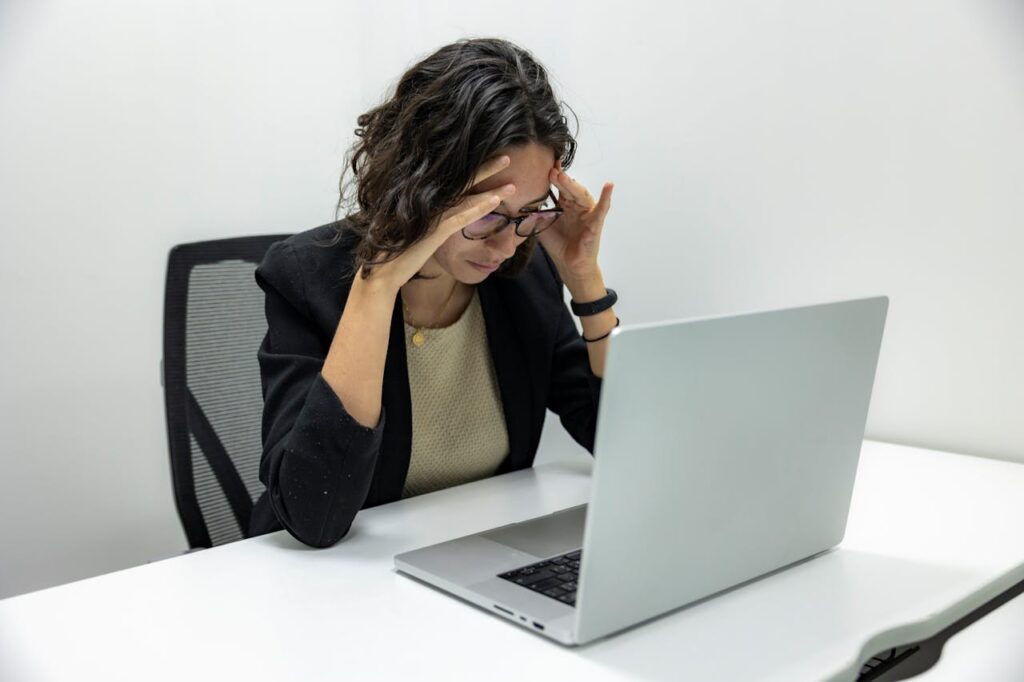 Femme assise au travail devant son ordinateur portable, elle a les mains sur les côtés du front, pensive - syndrome de l'imposteur - psychothérapie Paris 9 