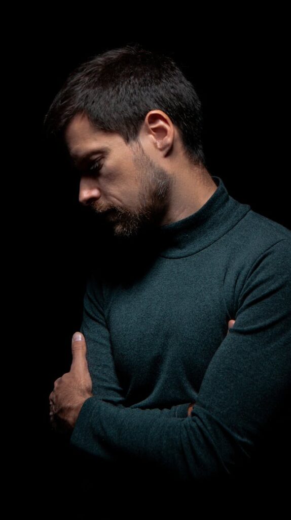 Homme sur fond noir, les bras croisés, pensif - syndrome de l'imposteur - psychothérapie Paris 9