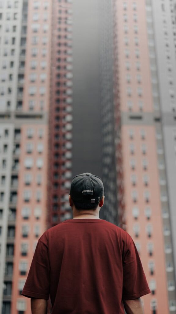 Homme en t-shirt rouge, avec une casquette, de dos, qui fait face à deux grandes tours d'immeuble, il est visiblement en réflexion, il doute - syndrome de l'imposteur - psychothérapie Paris 9