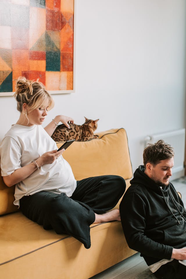 Femme enceinte assise sur le canapé et qui caresse son chat, utilise son téléphone pendant que son conjoint par terre est visiblement sur son ordinateur - IA et couple - psychothérapie Paris 9
