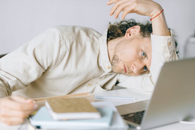 Homme le coude sur la table, le visage sur le bras, pensif et anxieux, devant son ordinateur et des documents en nombre - Procrastination - Psychothérapie Paris 9