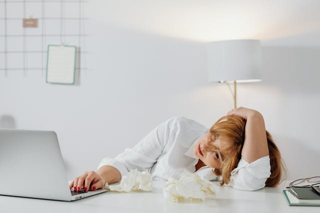 Femme qui semble dépassée par le travail, la main sur ses cheveux, le visage sur le bras, le coude sur son bureau, avec des papiers froissés sur la table, devant son ordinateur portable - Procrastination - psychothérapie Paris 9