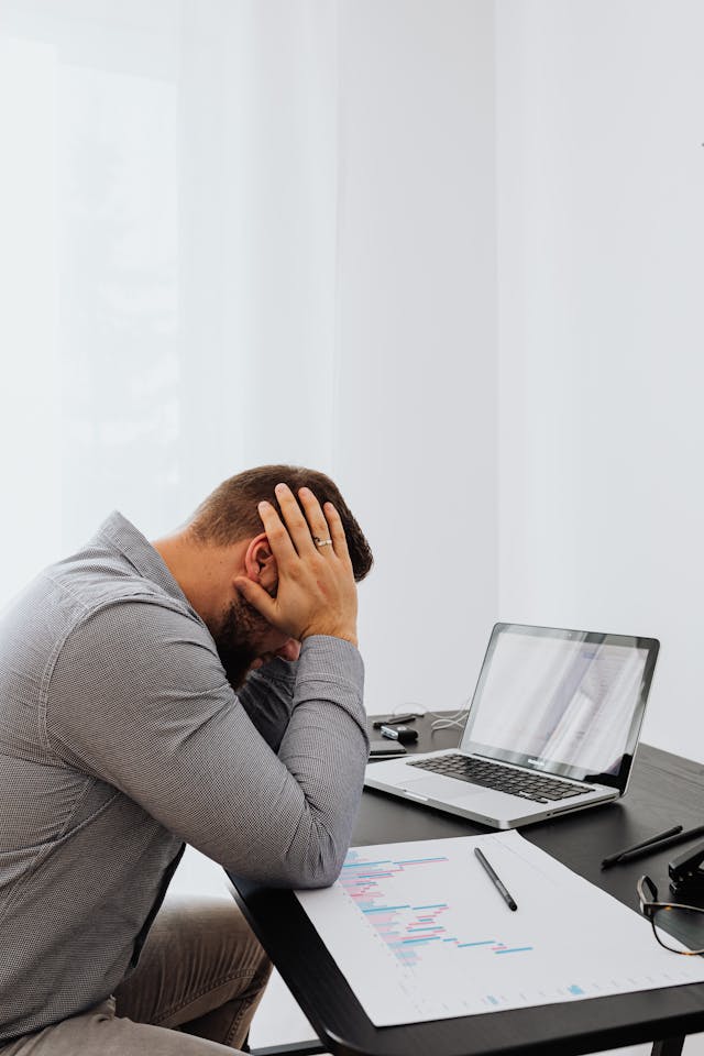 Homme tête baissée, les mains sur les tempes, qui semble harassé de travail devant son ordinateur - charge mentale - psychothérapie Paris 9