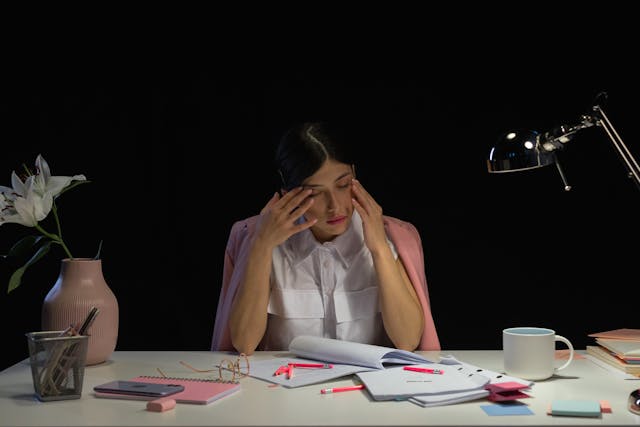 Femme les mains posées sur le visage, avec des feuilles et des surligneurs à son bureau, et qui semble dépassée et pensive - charge mentale - psychothérapie Paris 9
