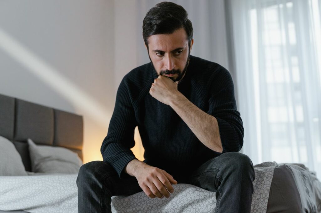 Homme assis sur son lit, qui se tient le menton avec sa main, pensif - pensée intrusive - psychothérapie Paris 9