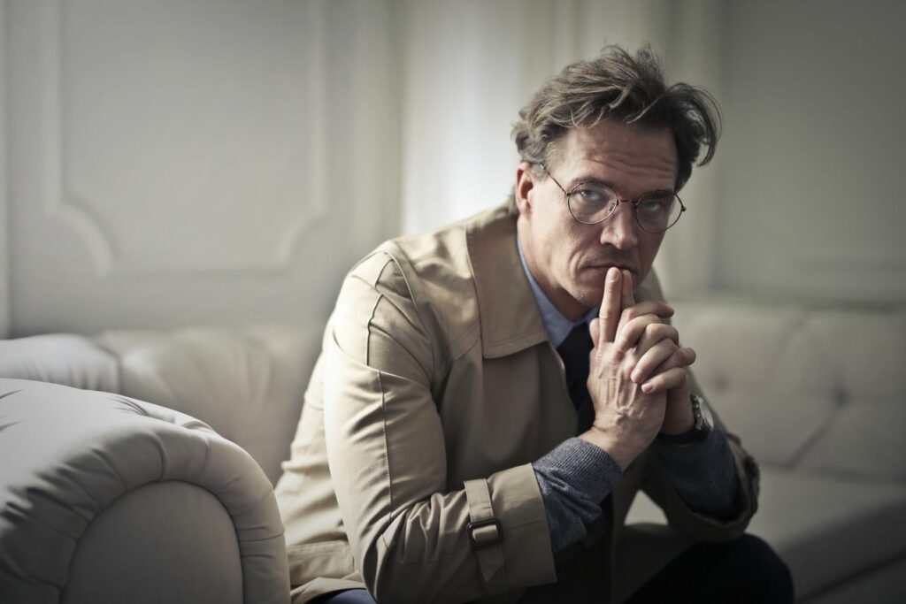 Homme assis sur un canapé, les mains et les index joints, pensif - pessimisme - Psychothérapie Paris 9