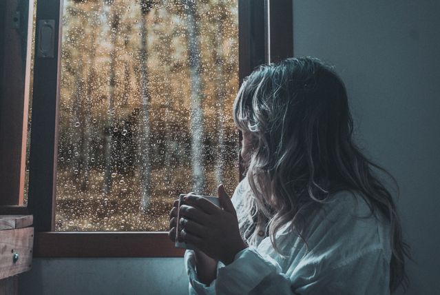 Femme qui tient sa tasse de boisson chaude et regarde, pensive, par la fenêtre, le paysage automnale - dysthymie - psychothérapie Paris