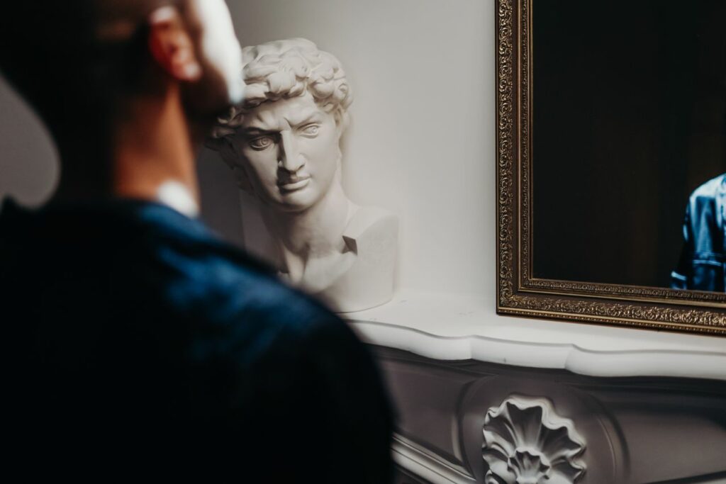 Un homme se regarde dans le miroir, une statue sur sa gauche fronce les sourcils - narcissisme - Psychothérapie Paris 9