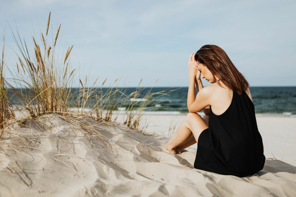 Une femme en robe noire est assise sur des dunes de sable près de l'océan, pensive - dépression estivale - psychothérapie Paris 9