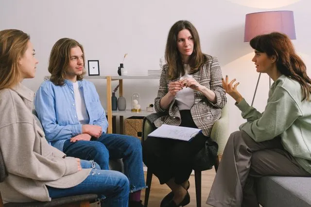 Thérapeute en face de trois adultes dans le cadre d'une thérapie familiale : une femme s'exprime avec un geste interrogatif de la main - thérapie contextuelle - Psychothérapie Paris 9 et Pantin