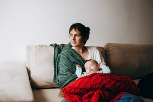 Femme qui tient son bébé sur ses genoux, assise sur le canapé, pensive - Troubles psychiques du post-partum