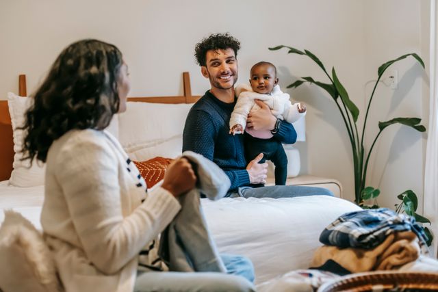 Un père porte son bébé, tous deux regardent leur mère avec le sourire pendant qu'elle plie le linge - Troubles psychiques du post-partum